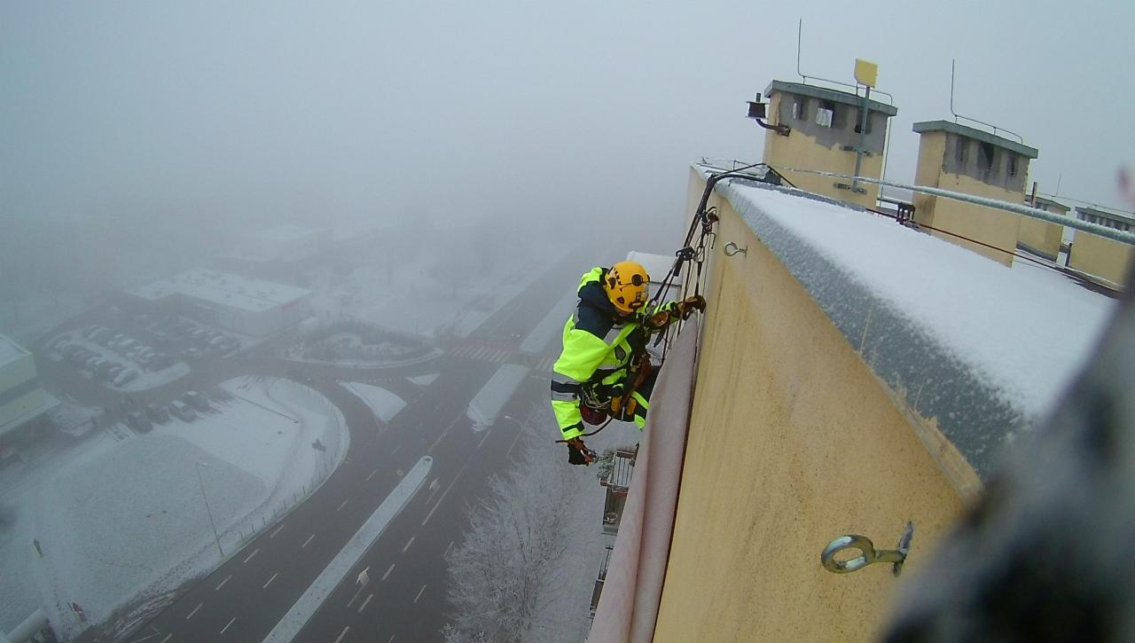 Pracownik w uprzęży, zabezpieczony linami, wykonuje prace na wysokości na ośnieżonej elewacji budynku w mglisty, zimowy dzień. Widok z góry na miasto spowite mgłą.