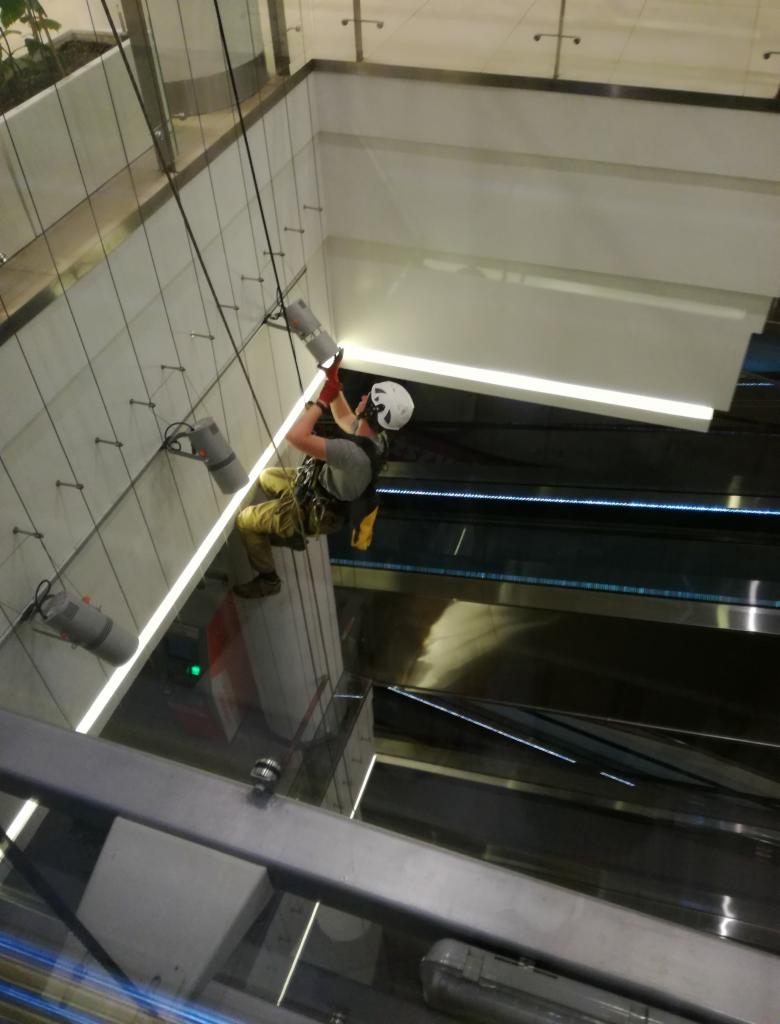 Pracownik w kasku i uprzęży, zawieszony na linach, czyści szklaną elewację wewnątrz budynku, używając specjalistycznego sprzętu. Widoczny fragment schodów ruchomych w tle.