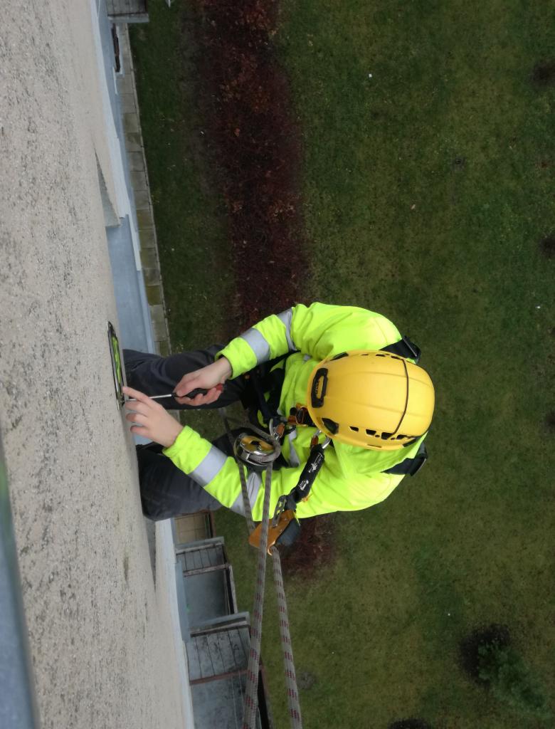 Pracownik w żółtym kasku i odblaskowej kurtce, zabezpieczony linami, przytwierdza element do elewacji budynku za pomocą śrubokręta, widok z góry na tle trawnika.