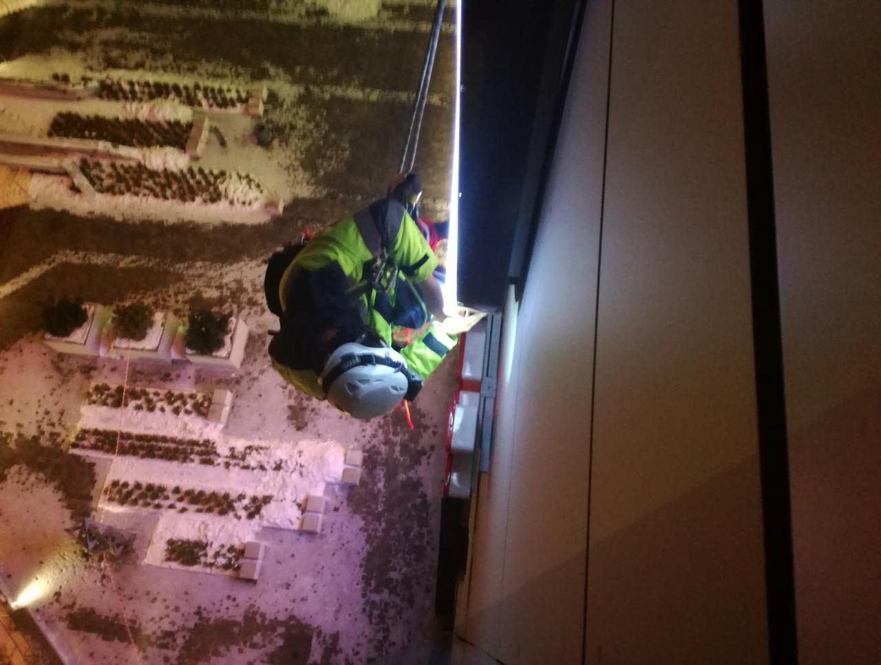 Pracownik na linach, w kasku i odblaskowej kurtce, wykonuje prace na wysokości na elewacji budynku, widok z góry na teren z elementami zieleni.