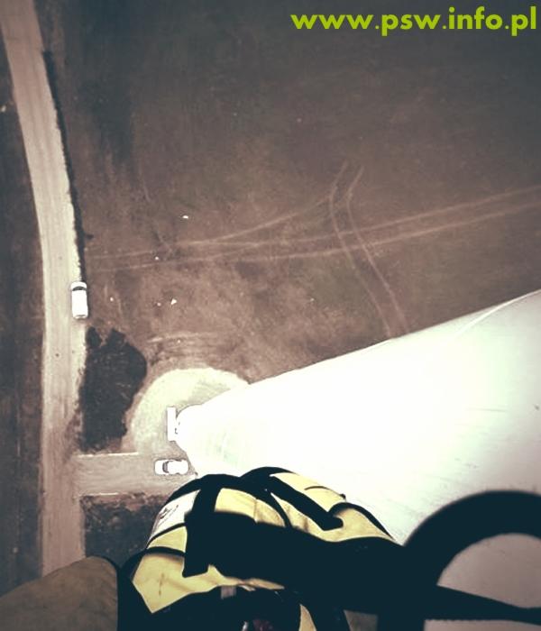 Widok z góry na białą elewację budynku w trakcie czyszczenia, z widocznymi samochodami na drodze i polach w tle, oraz fragmentem uprzęży pracownika na pierwszym planie.