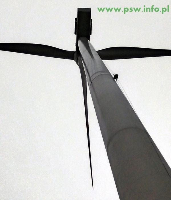 Czarno-białe ujęcie z dołu na turbinę wiatrową z widocznym pracownikiem na wysokości, w tle jasne niebo i adres strony www.psw.info.pl w prawym górnym rogu.