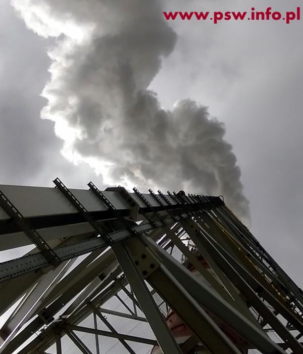 Metalowa konstrukcja wsporcza komina przemysłowego, z widocznym dymem unoszącym się na tle pochmurnego nieba, ujęcie z dołu.