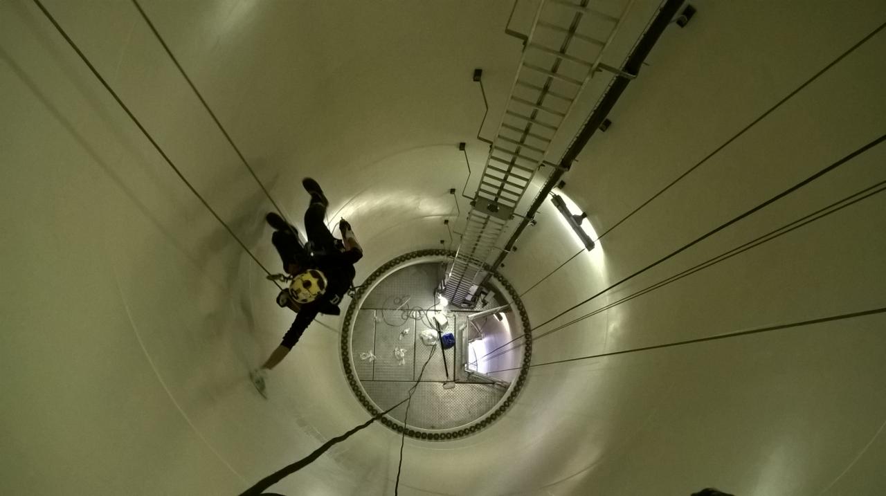 Pracownik w kombinezonie i kasku z linami asekuracyjnymi wewnątrz turbiny wiatrowej, widoczne drabiny i oświetlenie.