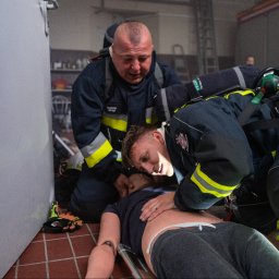 Sebastian Kazek EDUNAVIGATOR - Strażacy w kombinezonach ćwiczą resuscytację na manekinie w remizie. Widoczny w tle wóz strażacki i osoba filmująca. Marcin Drąg na kombinezonie.
