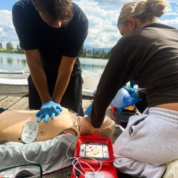 Sebastian Kazek EDUNAVIGATOR - Mężczyzna w rękawiczkach wykonuje RKO na manekinie, kobieta używa maski ambu. Obok otwarty defibrylator AED. Szkolenie pierwszej pomocy na drewnianym pomoście nad jeziorem.