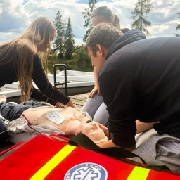 Sebastian Kazek EDUNAVIGATOR - Grupa osób ćwiczy resuscytację krążeniowo-oddechową na manekinie z defibrylatorem AED, na drewnianym pomoście nad wodą, obok czerwonej torby medycznej z emblematem.
