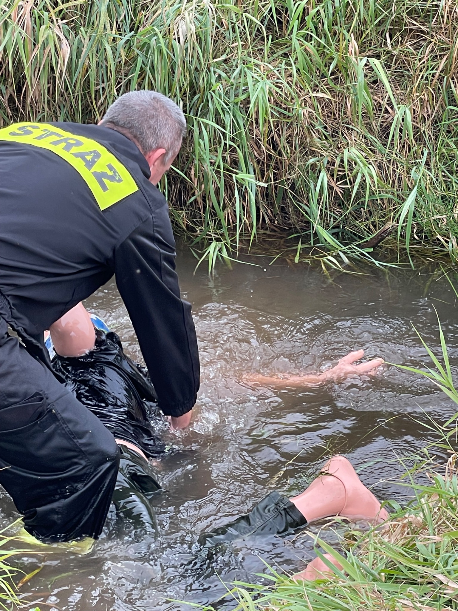 Akcja ratunkowa w Katowicach: strażak w czarnym uniformie z napisem 'STRAŻ' pomaga osobie tonącej w strumieniu, widoczne ręce i noga poszkodowanego wystające z wody.