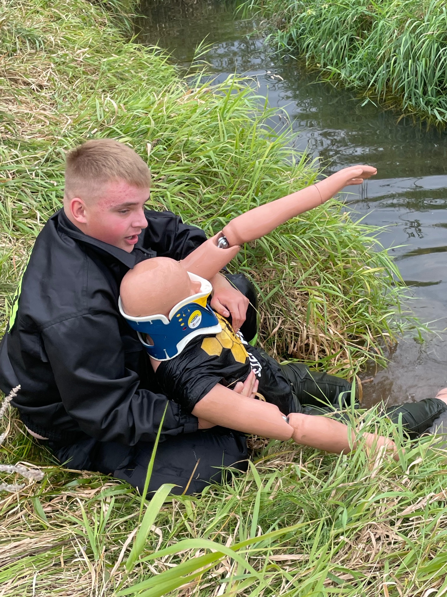 Młody mężczyzna w czarnej kurtce, siedząc w trawie nad brzegiem wody, udziela pomocy manekinowi z założonym kołnierzem ortopedycznym na szyi. Ramię manekina wystaje w kierunku wody.