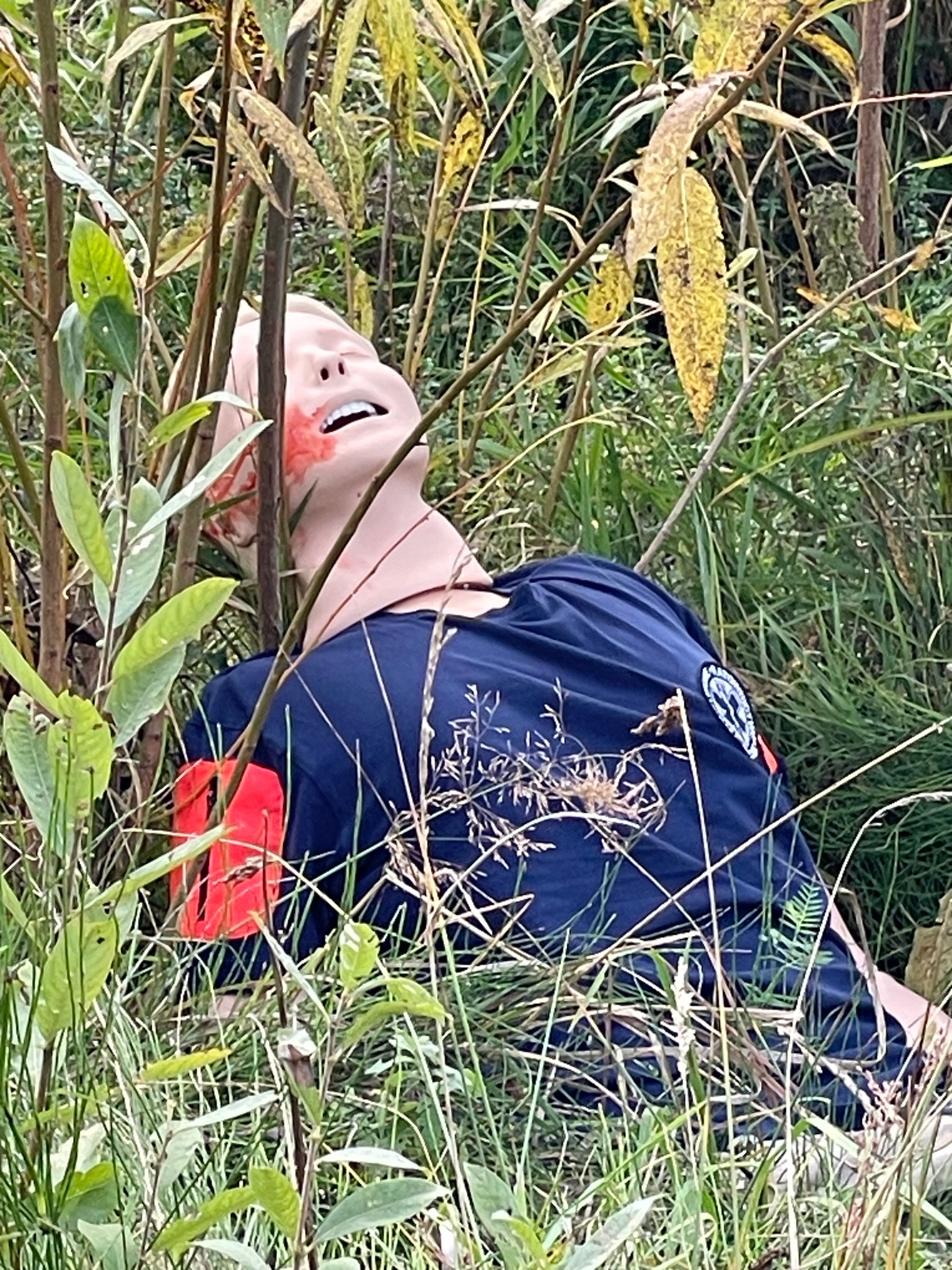 Manekin z symulowaną raną na twarzy, leżący w trawie podczas pozorowanego wypadku, widoczny fragment niebieskiej koszulki z naszywką