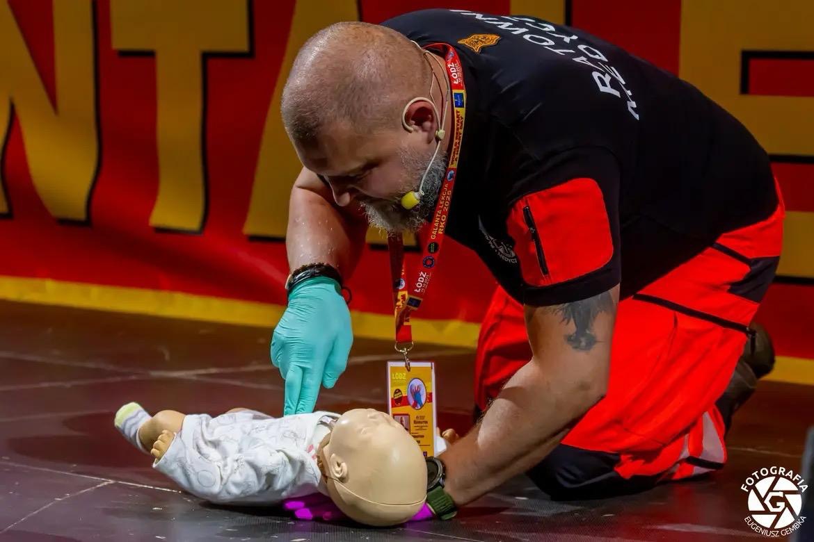 Instruktor w rękawicach, klęczący obok manekina niemowlęcia, demonstruje technikę resuscytacji podczas kursu pierwszej pomocy. Widoczny identyfikator 'Łódź'.