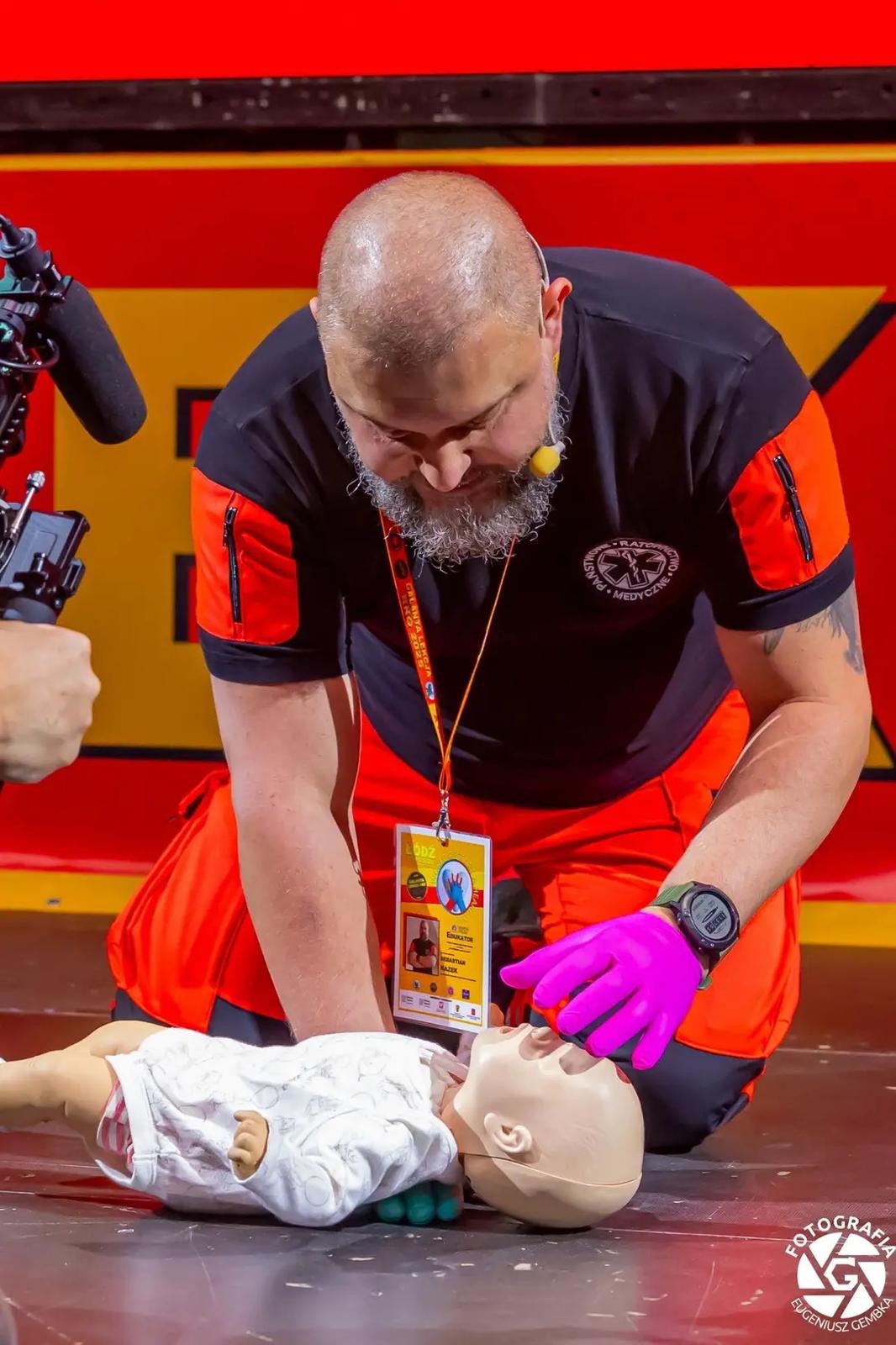 Instruktor z brodą w różowych rękawiczkach demonstruje resuscytację na manekinie dziecka. Widoczny identyfikator i mikrofon. Intensywne, sztuczne oświetlenie sceniczne.