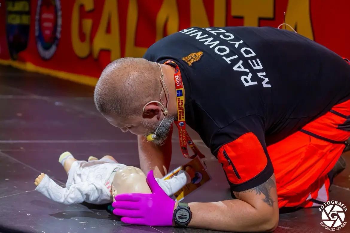 Instruktor z brodą w różowych rękawiczkach demonstruje resuscytację krążeniowo-oddechową na manekinie niemowlęcia podczas kursu pierwszej pomocy na scenie.