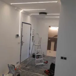 Wnętrze w trakcie remontu, z widocznymi listwami LED na suficie i ścianie, drabina, narzędzia i materiały budowlane rozłożone na podłodze zabezpieczonej folią.