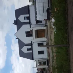 Nowy dom murowany z ciemnym dachem i białą elewacją, częściowo w rusztowaniach, widok z dołu na tle zachmurzonego nieba.