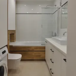 Nowoczesna łazienka z drewnianymi akcentami: zabudowana wanna z drewnianą obudową, biała umywalka nablatowa z szafką, toaleta wisząca obok pralki, beżowe płytki podłogowe, oświetlenie punktowe.