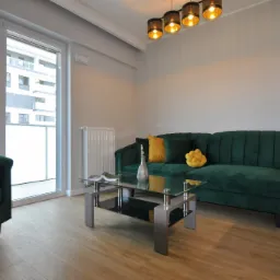 Jasne wnętrze salonu z drewnianą podłogą, szmaragdową sofą i fotelem, szklanym stolikiem kawowym, nowoczesną lampą wiszącą i dużym oknem balkonowym.