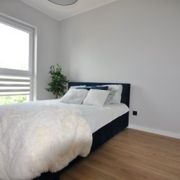 Rentsell Cengiz Yurekli - Jasny pokój z drewnianą podłogą, łóżkiem z ciemnym zagłówkiem, białymi poduszkami i narzutą, oraz rośliną w rogu, po remoncie, ściany pomalowane na szaro.