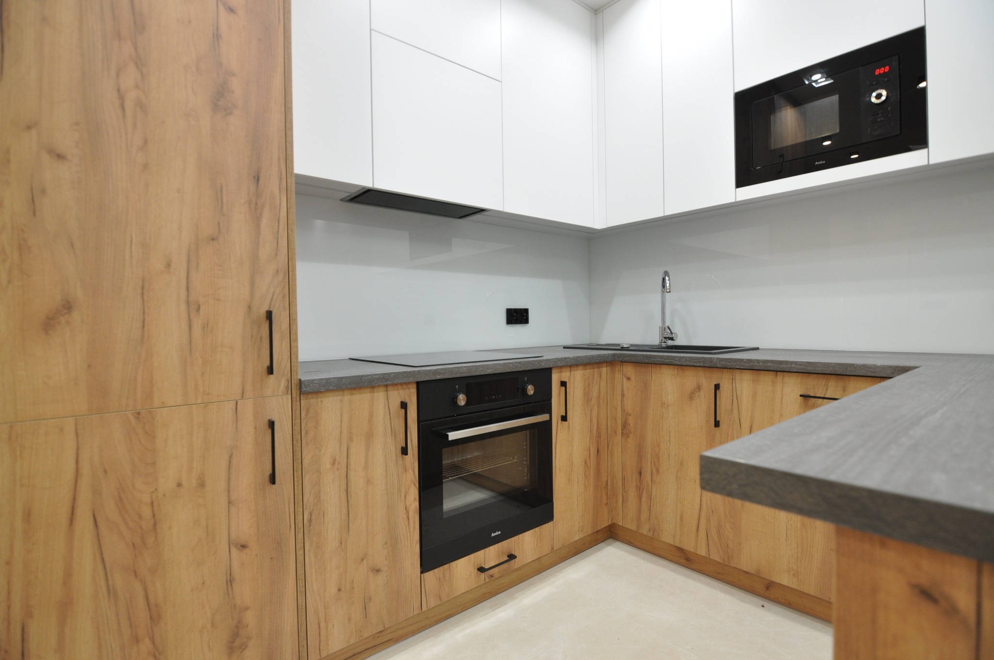 Nowoczesna kuchnia w odcieniach bieli i drewna, z zabudową kuchenną, szarym blatem, czarnym piekarnikiem i mikrofalówką, oraz minimalistyczną armaturą.