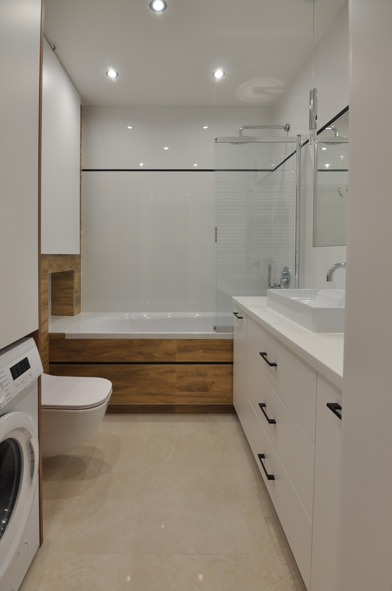 Nowoczesna łazienka z drewnianymi akcentami: zabudowana wanna z drewnianą obudową, biała umywalka nablatowa z szafką, toaleta wisząca obok pralki, beżowe płytki podłogowe, oświetlenie punktowe.