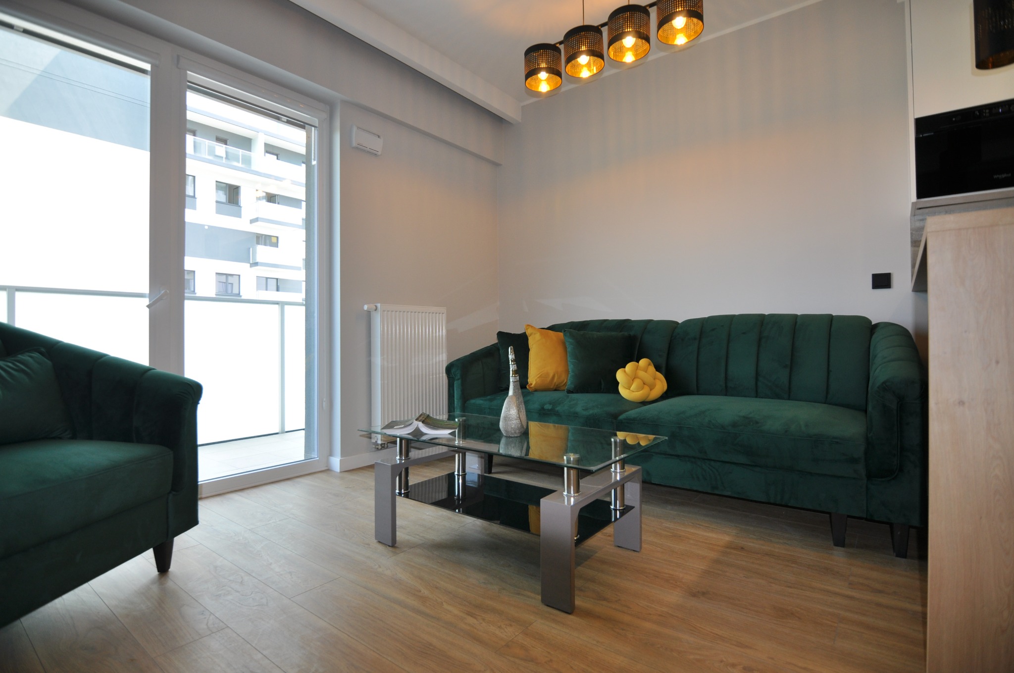 Jasne wnętrze salonu z drewnianą podłogą, szmaragdową sofą i fotelem, szklanym stolikiem kawowym, nowoczesną lampą wiszącą i dużym oknem balkonowym.