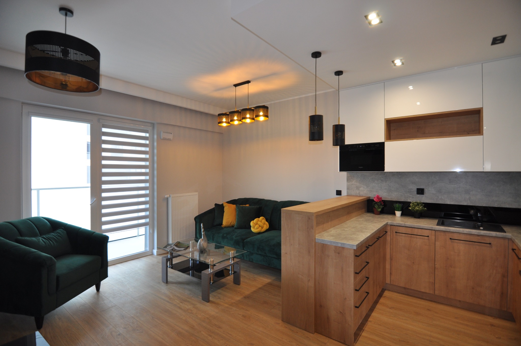 Nowoczesne wnętrze mieszkania z drewnianą podłogą, zieloną sofą i fotelem, biało-drewnianą zabudową kuchenną oraz dekoracyjnym oświetleniem, w tym wiszące lampy i rolety okienne.