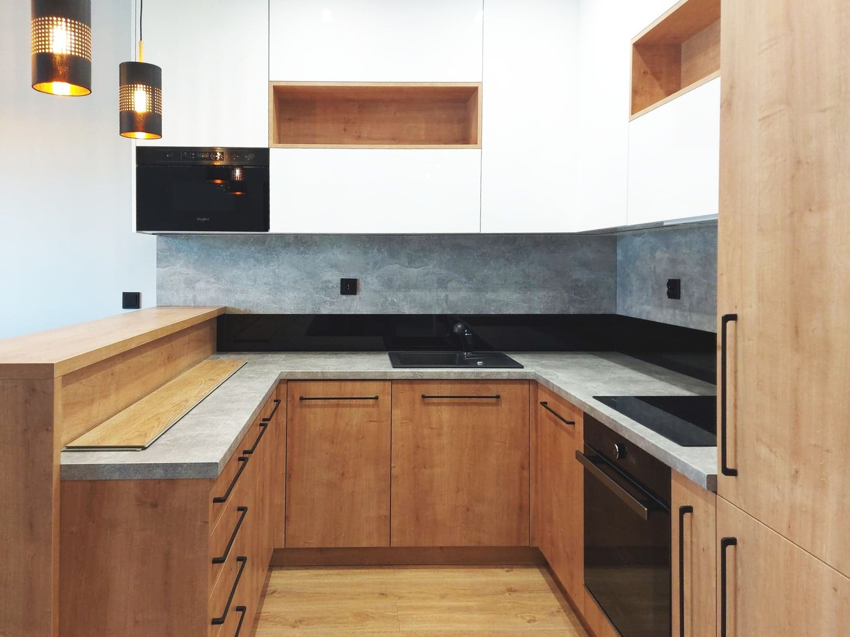 Nowoczesna kuchnia z drewnianymi szafkami o jasnym odcieniu, szarym blatem i betonową ścianą nad blatem, oświetlona wiszącymi lampami z ażurowymi kloszami.