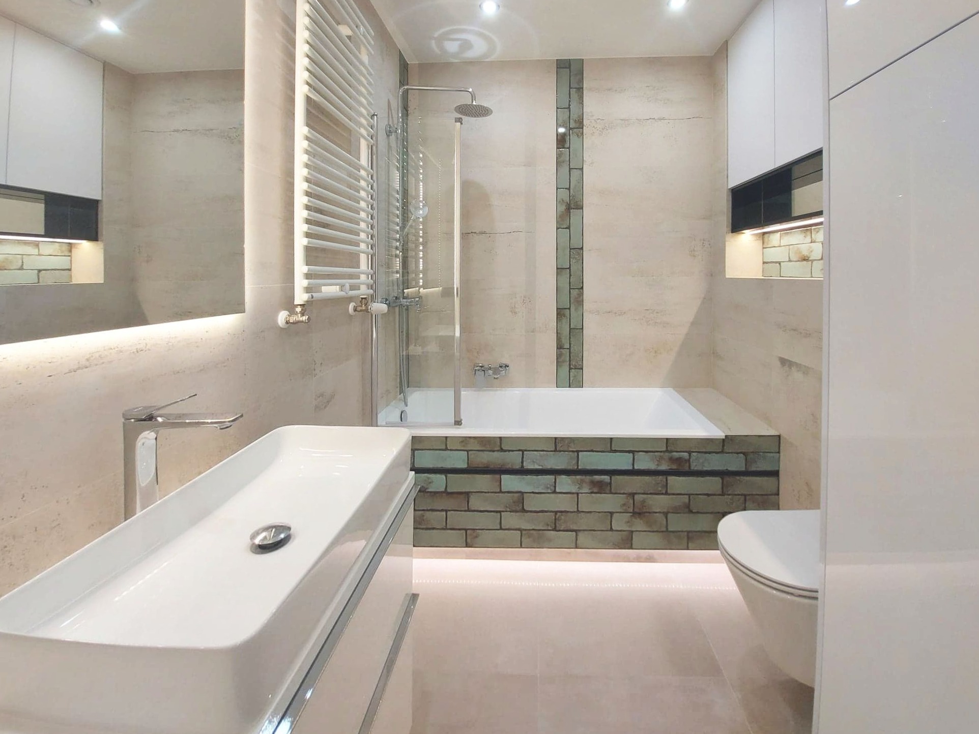 Nowoczesna łazienka z podświetlaną wanną obudowaną cegłą, minimalistyczną umywalką nablatową i grzejnikiem drabinkowym na ścianie z beżowych płytek.