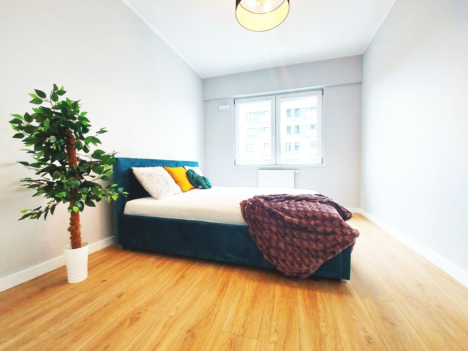 Jasna sypialnia z drewnianą podłogą, łóżkiem z turkusową ramą, białymi ścianami i sztuczną rośliną w białej doniczce