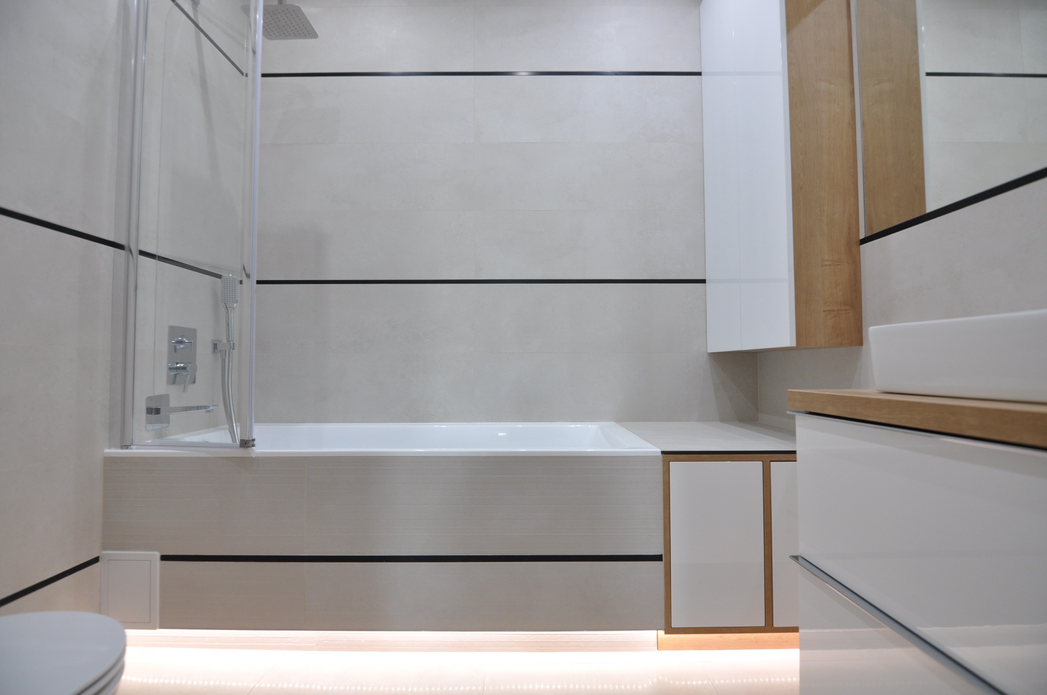 Wykończona łazienka z wanną obłożoną płytkami w odcieniach beżu z poziomymi czarnymi paskami, szklanym parawanem nawannowym i podświetleniem podłogowym LED, widoczna zabudowa meblowa z białymi...
