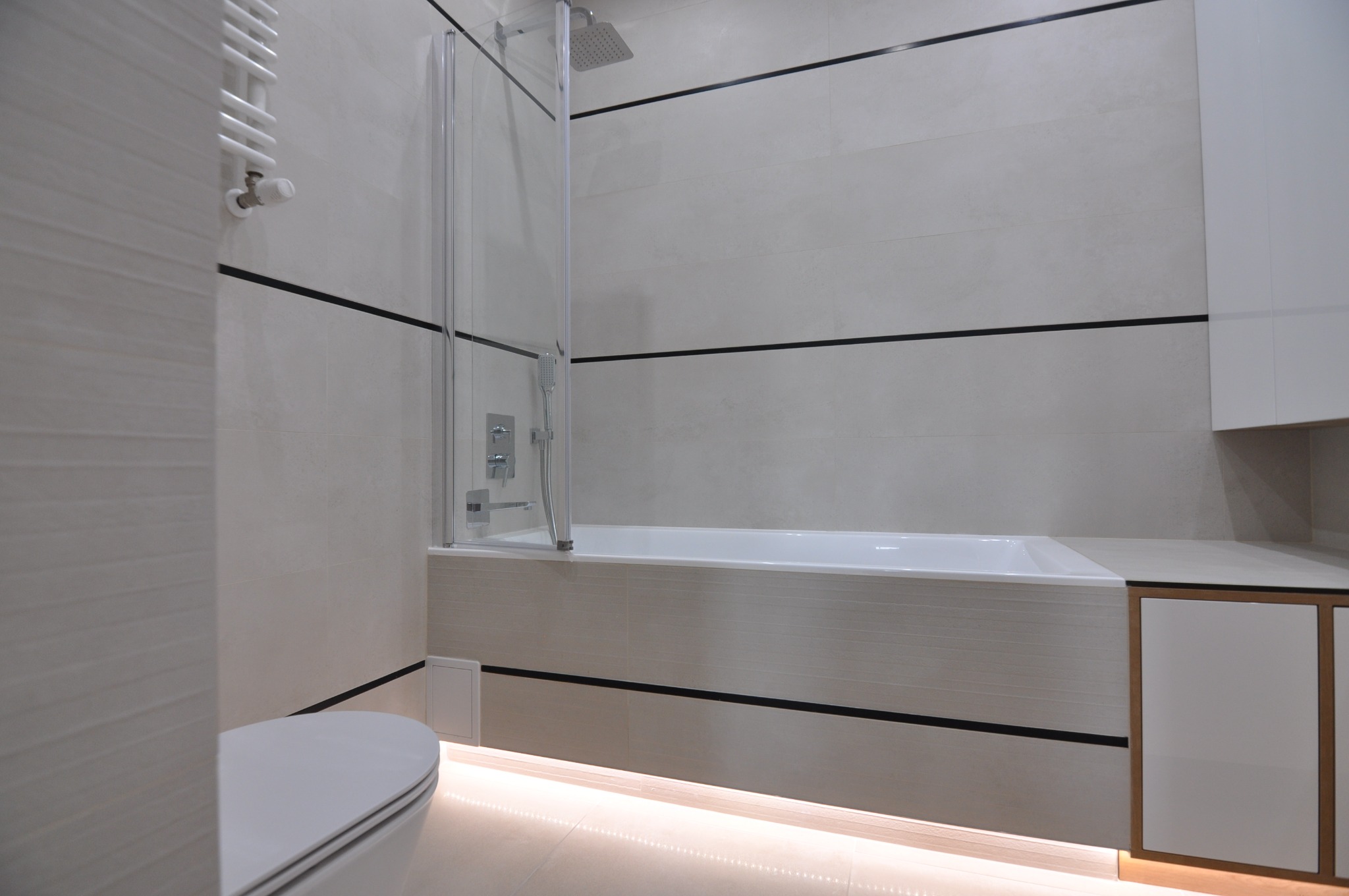 Nowoczesna łazienka z beżowymi płytkami, czarnymi akcentami i podświetlaną wanną za szklanym parawanem.