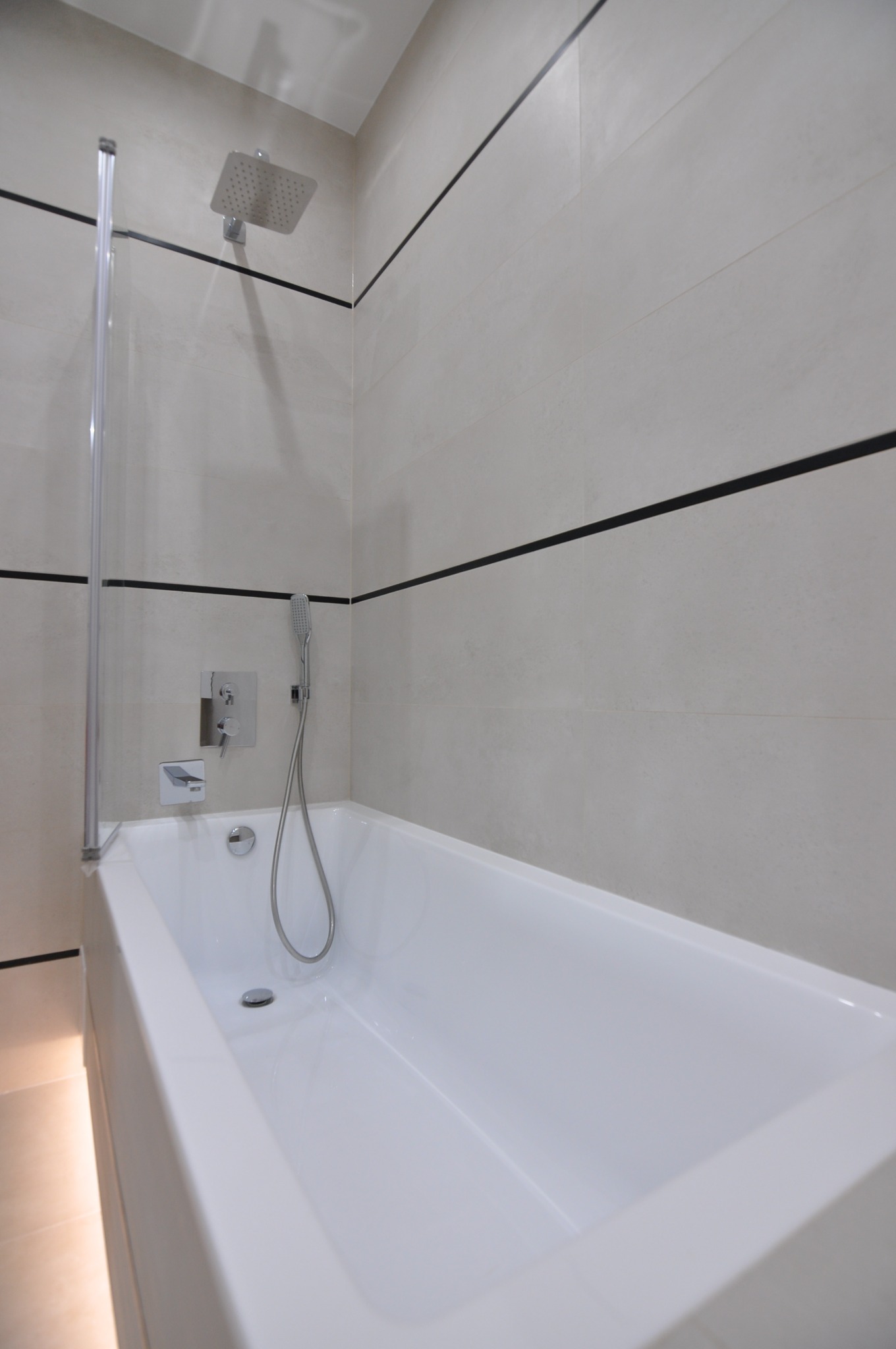 Nowoczesna, wykafelkowana łazienka z białą wanną, szklanym parawanem, deszczownicą i baterią podtynkową, akcentowana czarnymi pasami na jasnych płytkach.