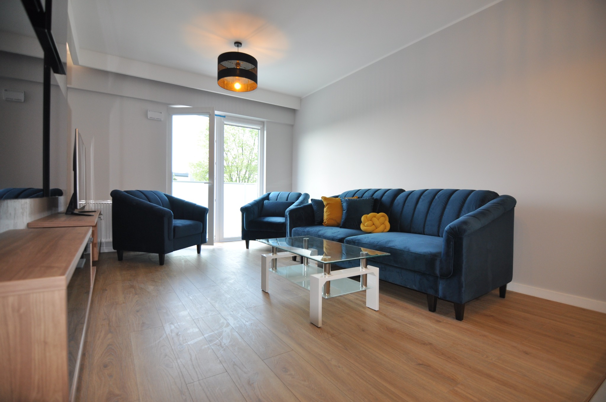 Salon z drewnianą podłogą, niebieską sofą i fotelami, szklanym stolikiem kawowym i nowoczesnym oświetleniem, świeżo po remoncie.