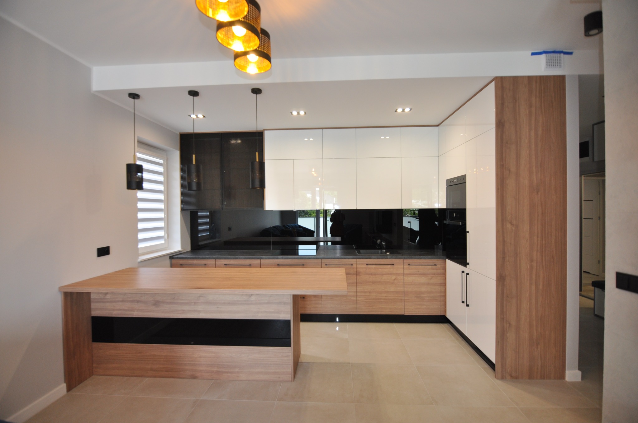 Nowoczesna kuchnia z białymi i drewnianymi szafkami, czarnym blatem i wyspą z elementami w kolorze drewna i czerni, oświetlona wiszącymi lampami z abażurami w kolorze złota.