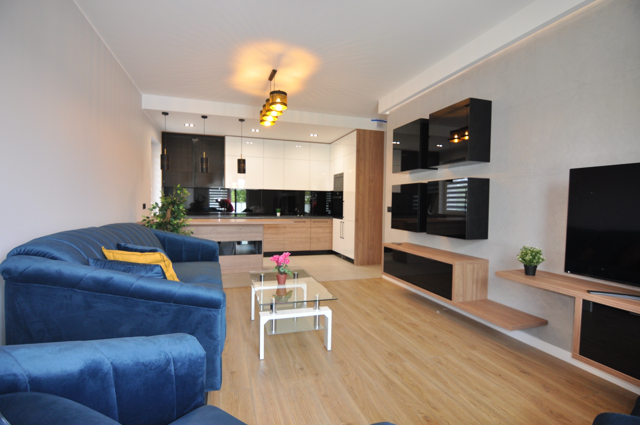 Jasne wnętrze salonu z aneksem kuchennym, ściana wykończona gładzią, podłoga z paneli drewnopodobnych, nowoczesne meble.