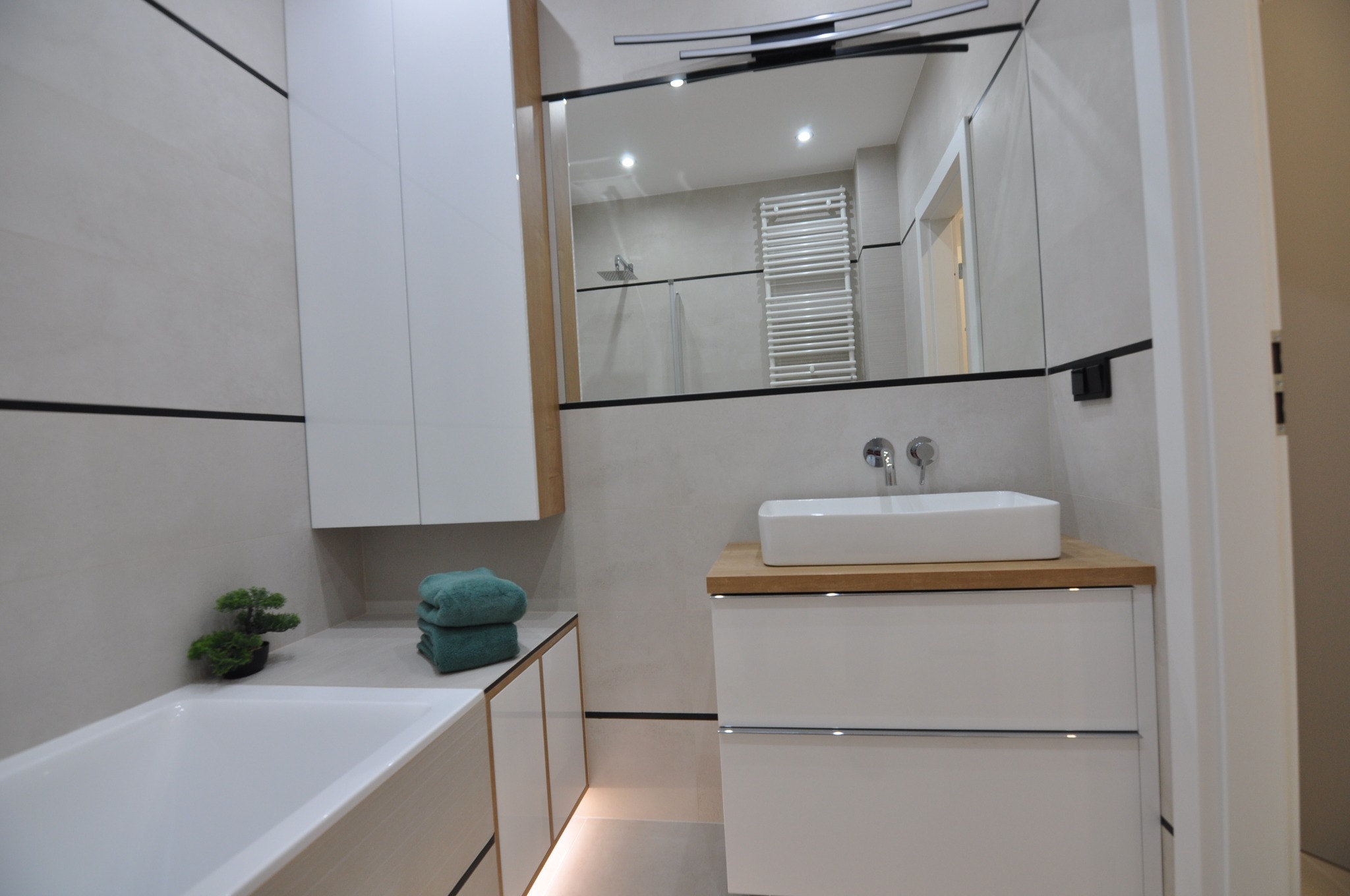 Nowoczesna łazienka z białymi meblami, prostokątną umywalką nablatową na blacie z jasnego drewna i lustrem z oświetleniem, widoczne elementy wykończenia ścian i armatury.