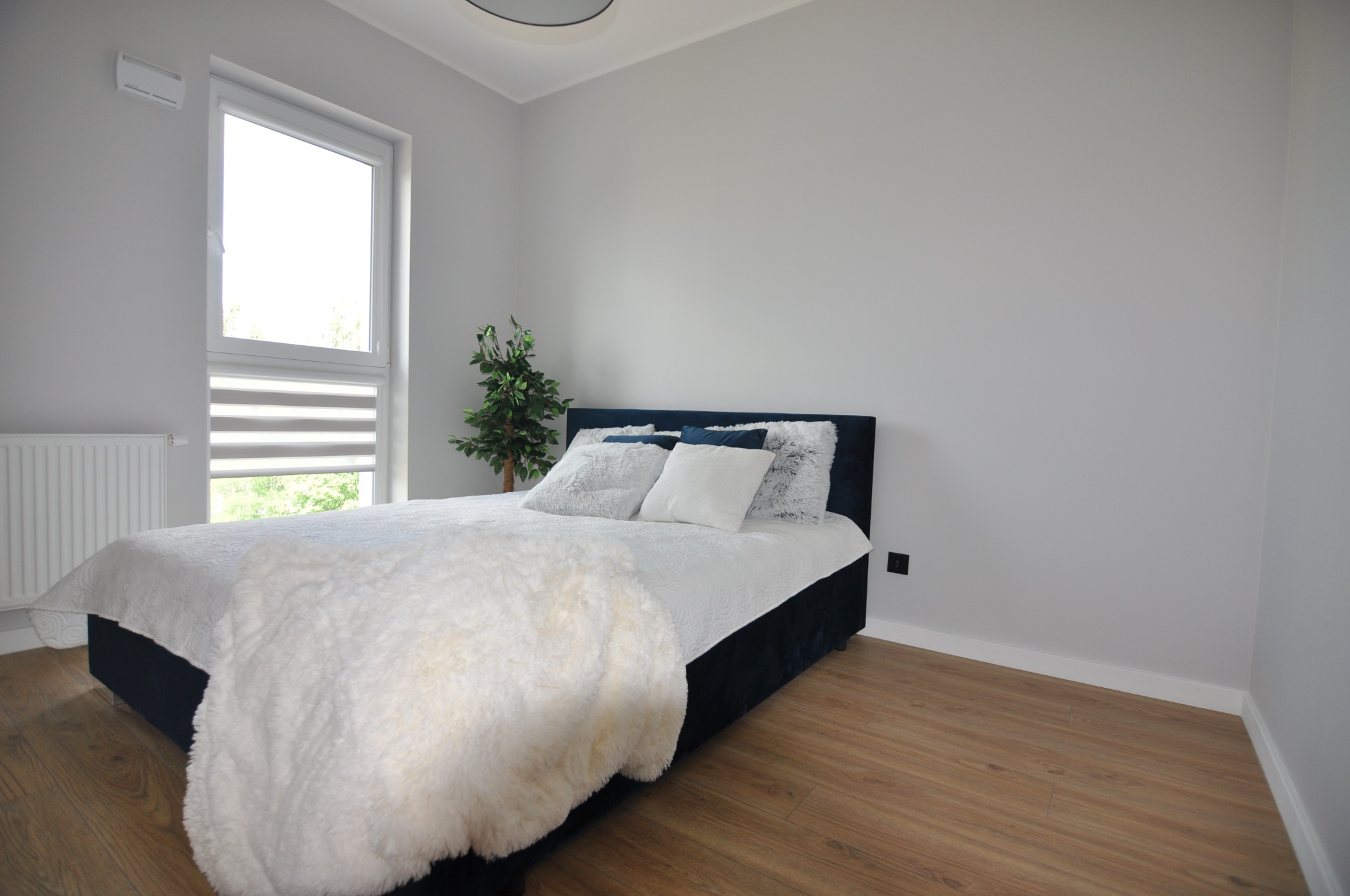 Jasny pokój z drewnianą podłogą, łóżkiem z ciemnym zagłówkiem, białymi poduszkami i narzutą, oraz rośliną w rogu, po remoncie, ściany pomalowane na szaro.