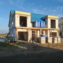 Niedokończony dwupiętrowy budynek z białych bloków, z niebieską folią na dachu, palety z cegłami Roben leżące przed budynkiem, widok z dołu na tle błękitnego nieba.