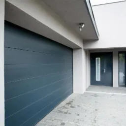 Nowoczesna, pozioma brama garażowa w kolorze grafitowym, z subtelnymi przetłoczeniami, na tle jasnoszarej elewacji domu z wejściem i małym drzewkiem w drewnianej donicy.