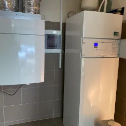 Białe urządzenie grzewcze marki Vaillant z otwartymi drzwiczkami panelu sterowania, widoczne srebrne rury wentylacyjne i szara płytka podłogowa.