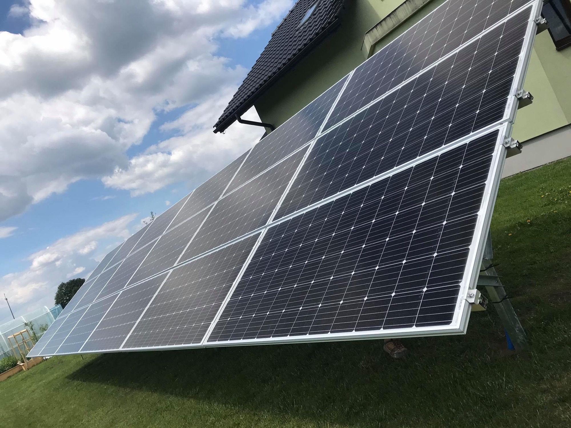 Panel słoneczny ustawiony na trawniku przed domem z zieloną elewacją i ciemnym dachem, widoczne niebieskie rury przy podstawie konstrukcji.