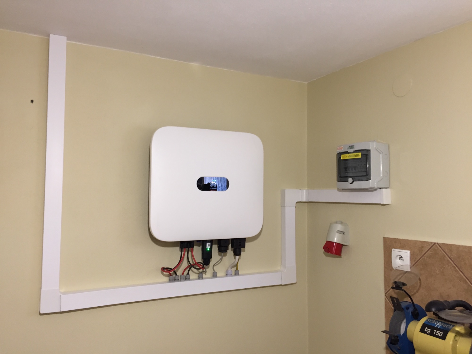 Biały falownik fotowoltaiczny marki Huawei zamontowany na ścianie z widocznymi przewodami i zabezpieczeniami, obok skrzynka elektryczna i gniazdo siłowe.