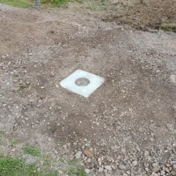 Betonowa studzienka rewizyjna z okrągłym otworem, umieszczona w ziemi pokrytej drobnymi kamieniami, z fragmentem trawy i butem w tle.