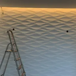 Falista, biała ściana dekoracyjna podświetlona od góry, z widoczną drabiną i wystającymi kablami elektrycznymi.