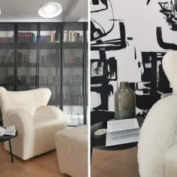 Kremowy fotel typu teddy z podnóżkiem na parkiecie w jodełkę, obok czarny stolik z wazonem i otwartą książką, na ścianie abstrakcyjny czarno-biały wzór, w tle przeszklona biblioteczka.