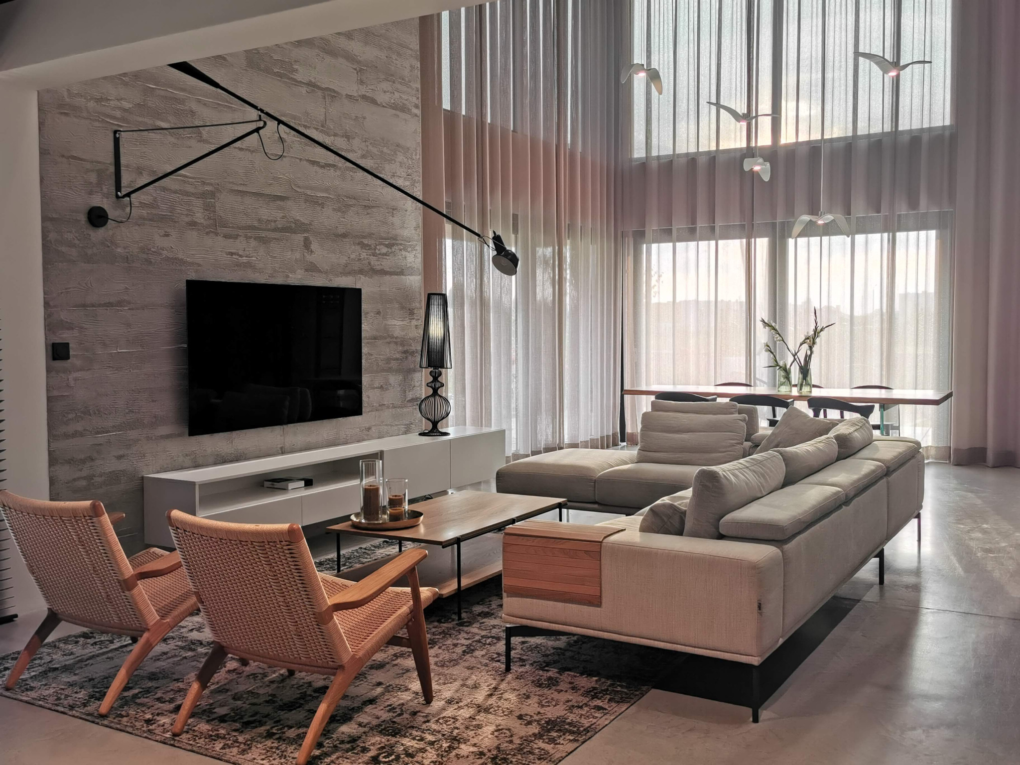 Jasne, przestronne wnętrze salonu z betonową ścianą akcentującą, minimalistyczną lampą na wysięgniku, szarym narożnikiem z drewnianym akcentem, dwoma wiklinowymi fotelami i dekoracyjnymi lampami...