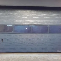 Szara segmentowa brama garażowa z czterema prostokątnymi oknami w górnej części, widoczny zamek i logo producenta u dołu, zamontowana w surowym otworze z widocznymi elementami konstrukcyjnymi.