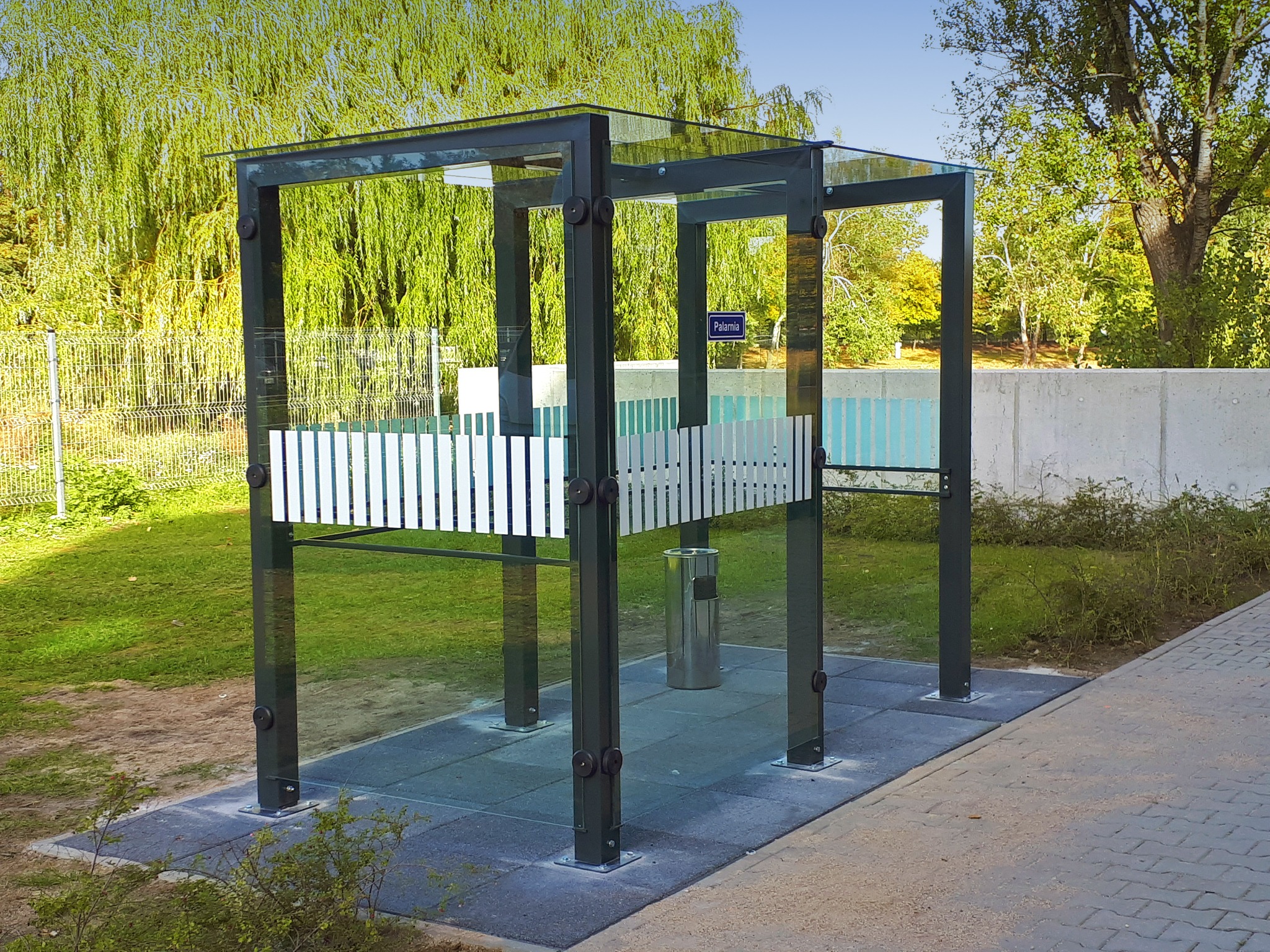 Nowoczesna wiata palarnia z metalową konstrukcją, szklanym dachem i częściowo ażurowymi ściankami, umieszczona na zewnątrz na kostce brukowej, z widocznym koszem na śmieci i tabliczką informacyjną.