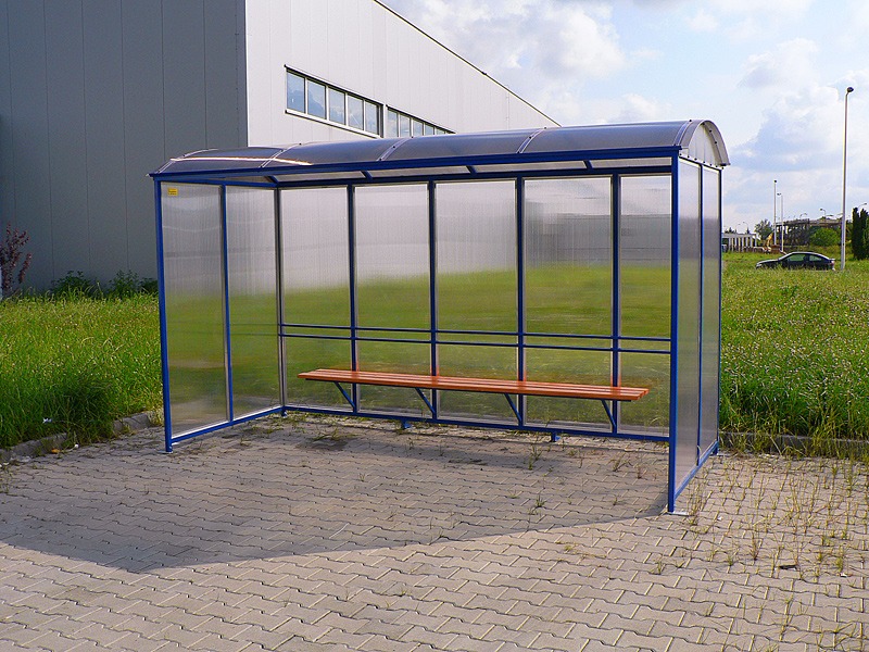 Zadaszona wiata przystankowa z niebieską ramą i drewnianą ławką, ustawiona na brukowanym placu, z widokiem na budynek przemysłowy i zieleń w tle.