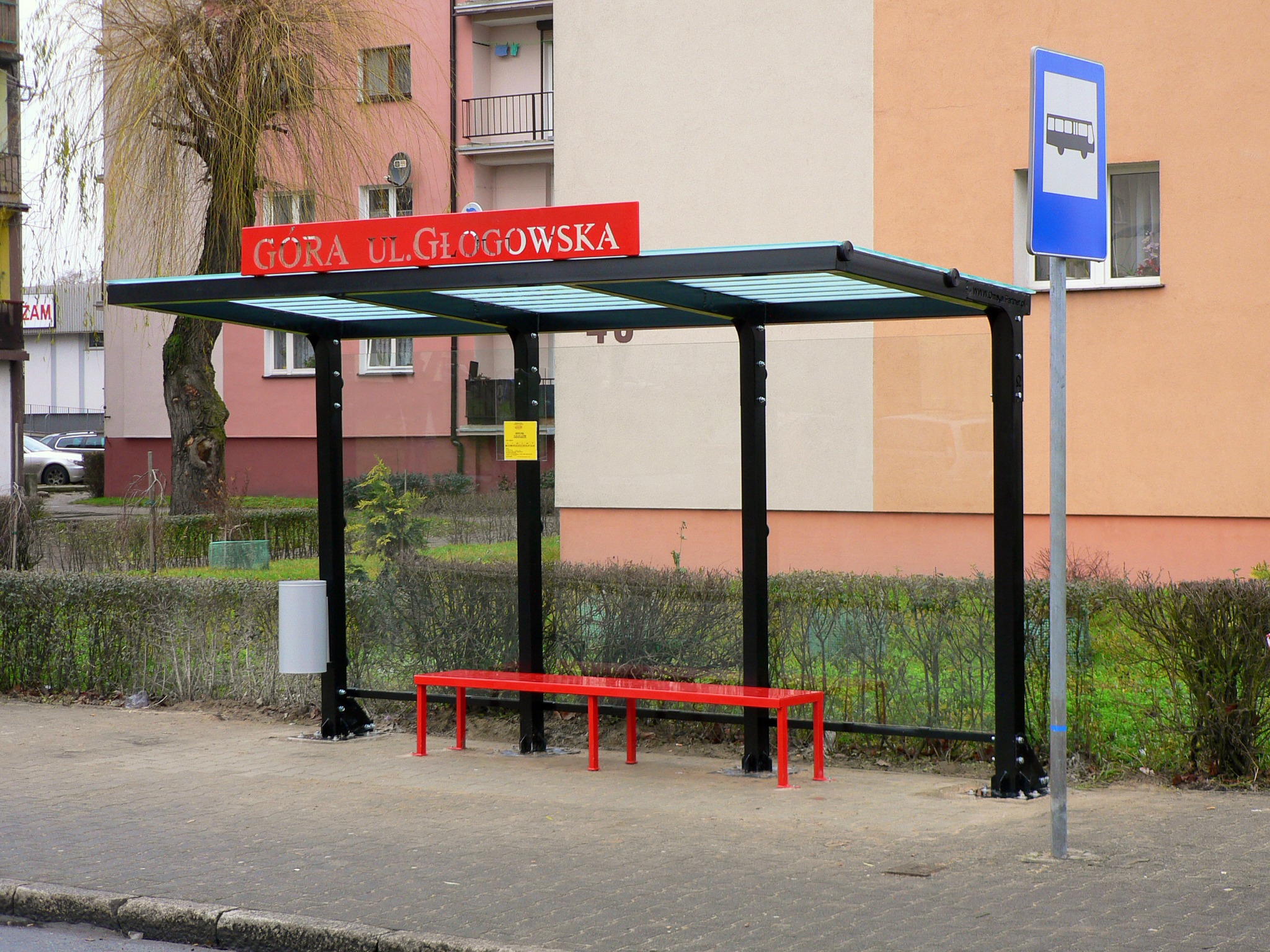 Przystanek autobusowy z czerwoną ławką i tablicą z nazwą ulicy 'Góra Ul. Głogowska', na tle budynków mieszkalnych i żywopłotu.